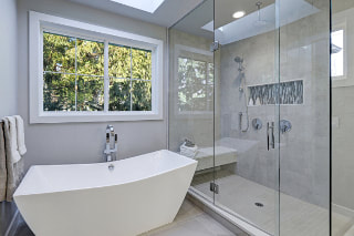 Picture of gray bathroom with white modern tub and large free staning shower enclosed in glass. large window across back of tub with white trim. 
