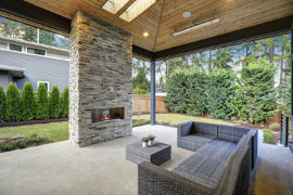 Image of a wood panel ceiling covered porch with a gray and white moder brick fireplace. Gray couch and matching coffee table. Concrete flooring ties the entire porch together.
