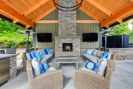 Picture of a back porch with a cedar wood ceiling. Gray floor to ceiling stone firepace and outdoor kitchen. Two couches and two chairs of natural fibers facing each other. 