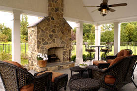  Picture of a outdoor living room under a covered porch with a tan stone fireplace. Dark brown furniture and ceiling fan. 