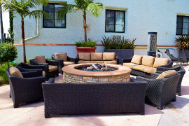 Photo of a patio with dark brown furniture facing a large stone round fire pit. Surrounded by palm trees and greenery.