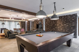 Picture of newly remodeled basement with white wood floors and dark brick accent wall. Dark wood pool table in the center with gray felt covering. Gray pendant lighting accenting pool table.