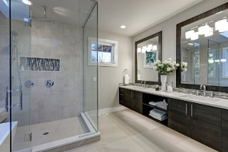 Picture of country french bath with large gray tile standup shower. Flat dark wood cabinets and mirror trim that sit over light counters and sinks. Light gray floor tiles with recessed lighting.