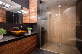 Picture of a modern washroom with black counters and grained wood cabinets. Large tan tile standup shower with rain shower head.