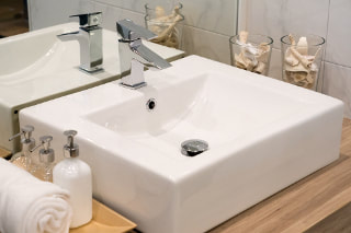 White ceramic square modern sink sitting on a wood counter with stainless steel faucets.