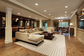 Picture of a renovated basemet with oak floors surrounding white tile square accent rug. Tan L-shaped couch and large dark wood bar along wall. All furniture is dark traditional wood. Recessed lighting throughout the room.