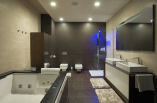 Large master bathroom photo with jetted white tub and sinks. Modern gray flooring and accent wall with white accent rugs. Recessed lighting with light walls.