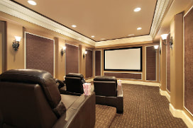 Picture of a home theater with tan and dark tan panels. Leather chairs staggared infront of large theater screen. Dark brown texture carpeting. Light tan ceiling with recessed lighing.